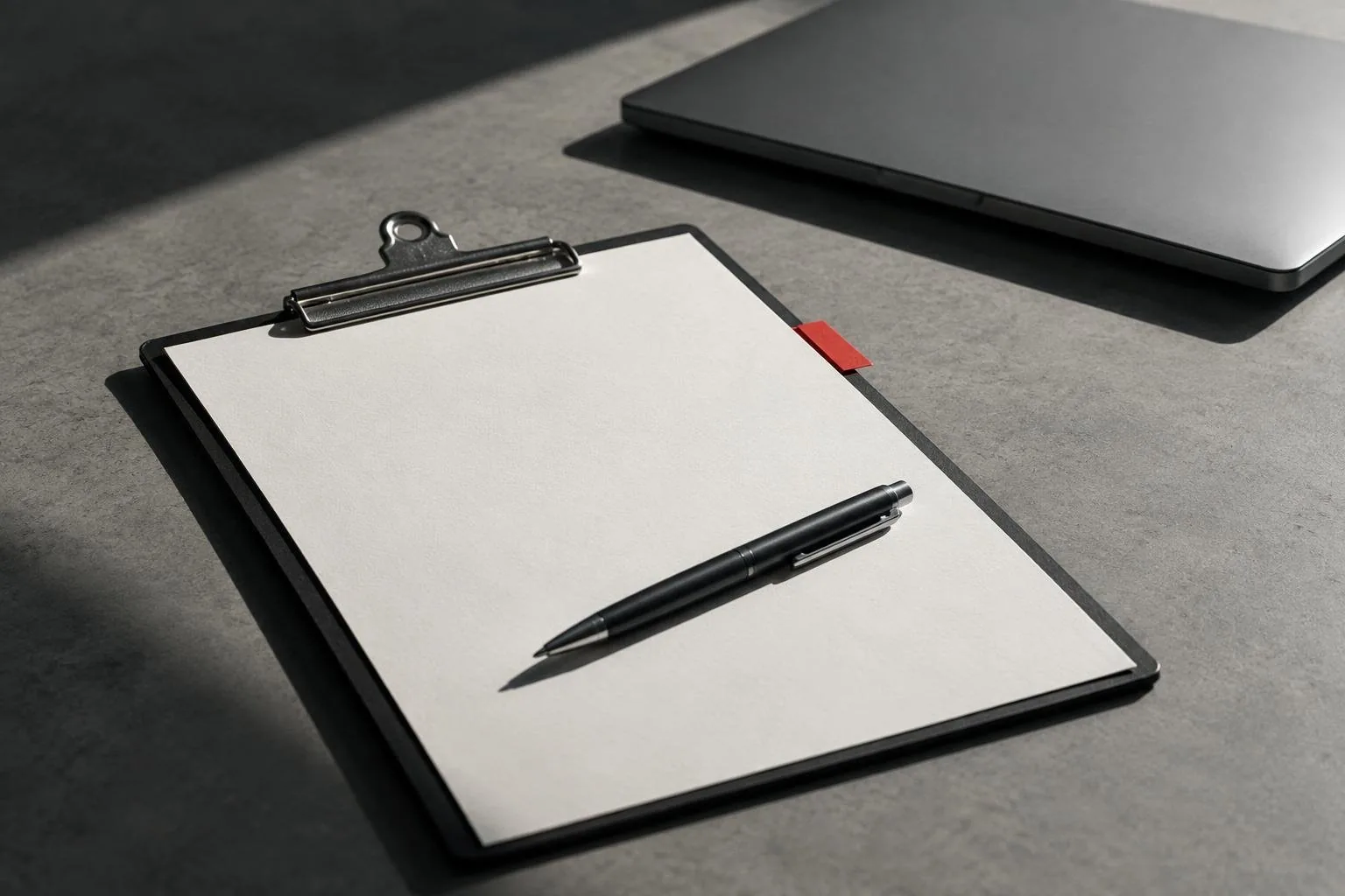 A clipboard with a blank quote sheet, a red tab on its edge and a pen laid across it, a closed laptop behind